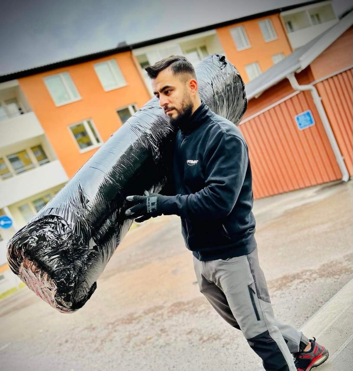 En flyttare från Optimus i svart arbetsjacka bär en inplastad rulle på axeln utanför ett flerfamiljshus